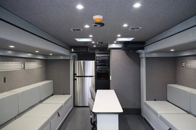 An interior image of a rehabilitation vehicle kitchen with a full fridge, storage, table, chairs and bench seating. 