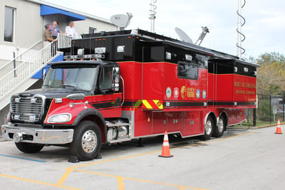 A red mobile command vehicle with a black top is parked outside a building with towers raised and orange cones at the front and rear of the vehicle. 