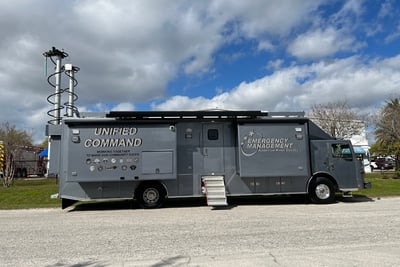 A grey mobile command vehicle is parked showing the raised antenna, with the slide out sections and stairs fully deployed. 